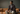 Model sitting on a burnt orange couch with cream pants, checkered jacket and a cigar in his hands