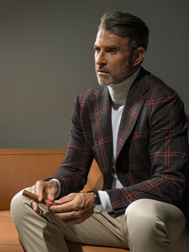 Model sitting on a burnt orange couch with cream pants, checkered jacket and a cigar in his hands